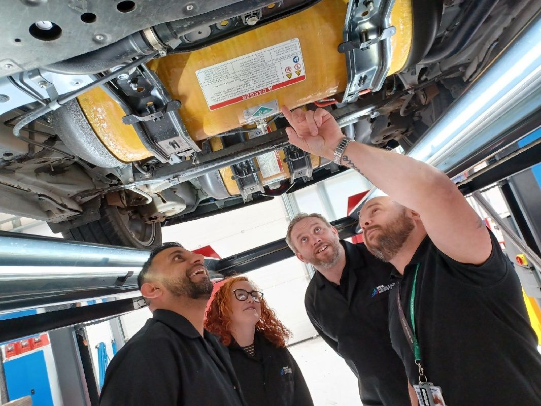 Group of adults, 1 asian male, 1 white female and two white males looking at the underside of a car