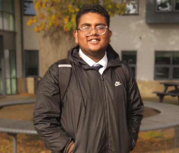 A young man smiles outdoors in a black jacket, with autumn leaves behind him.