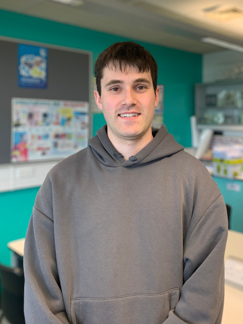 Tom wearing a grey hoodie stood in a classroom with posters